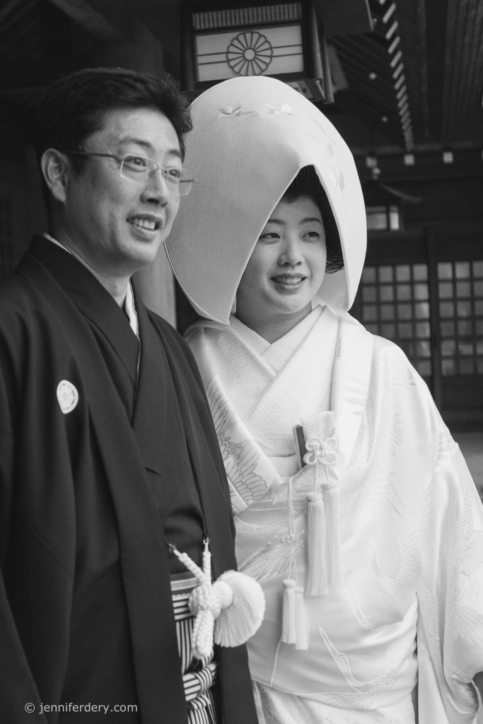 A smiling couple in traditional Japanese wedding attire; the bride wears a white kimono and a large headpiece, while the groom wears a black kimono. The photo is in black and white and appears to be taken at a temple.