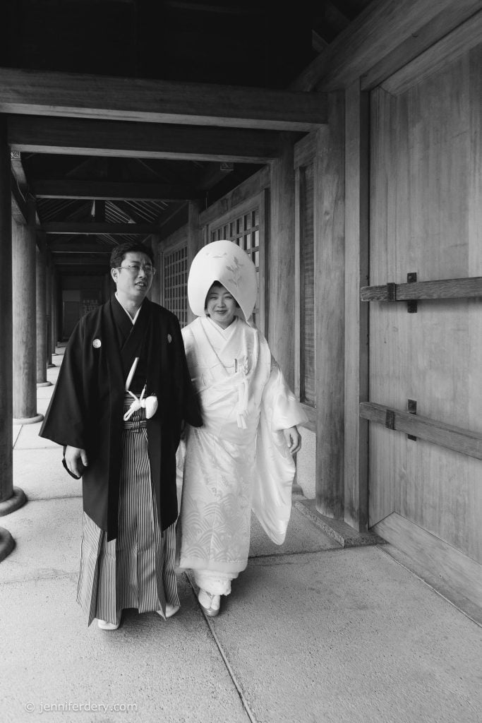 A couple in traditional Japanese wedding attire walks together in a wooden corridor. The groom wears a black kimono, and the bride wears a white kimono with a large, round headpiece. The atmosphere is serene and joyful.