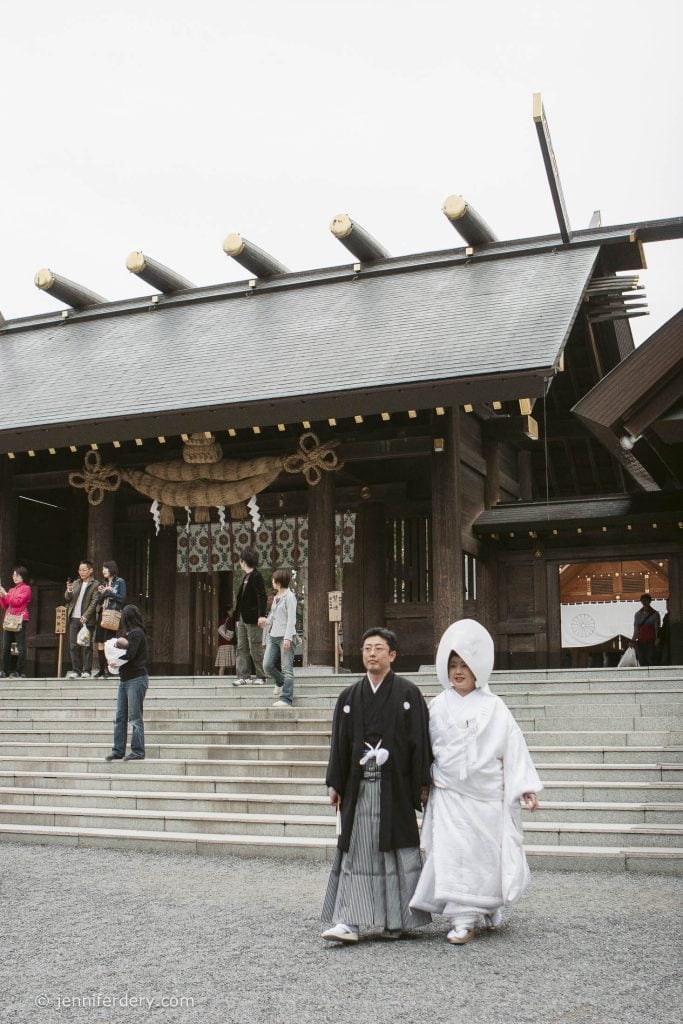 A couple in traditional Japanese wedding attire stands in front of a Shinto shrine. The groom wears a black kimono and hakama, while the bride wears a white kimono and a large headpiece. People are seen on the shrine steps behind them.