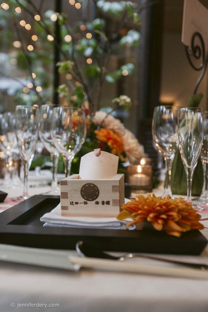 A beautifully set table with wine glasses, orange flowers, and a wooden Japanese sake box holding a rolled napkin, under soft, warm lighting and blurred, festive bokeh in the background.