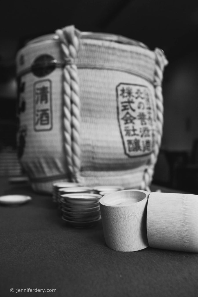 A large traditional Japanese sake barrel sits in the background, with a stack of small wooden sake cups on a table in the foreground. The image is in black and white.