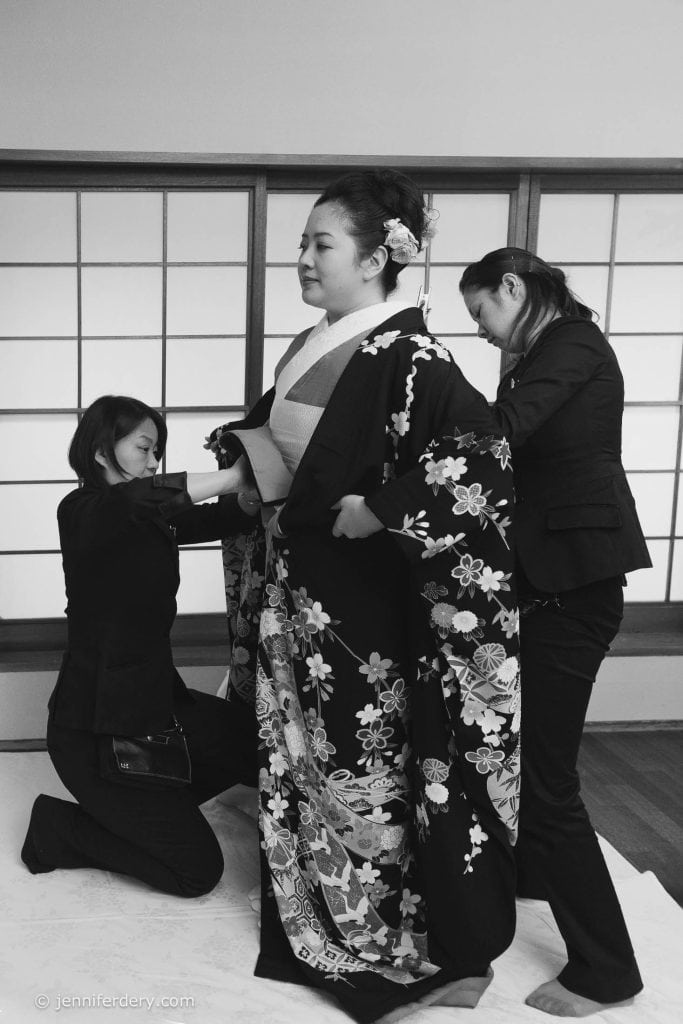 A woman stands in a traditional kimono with floral patterns, while two women help adjust her outfit. The scene takes place indoors in front of shoji screens. The image is in black and white.