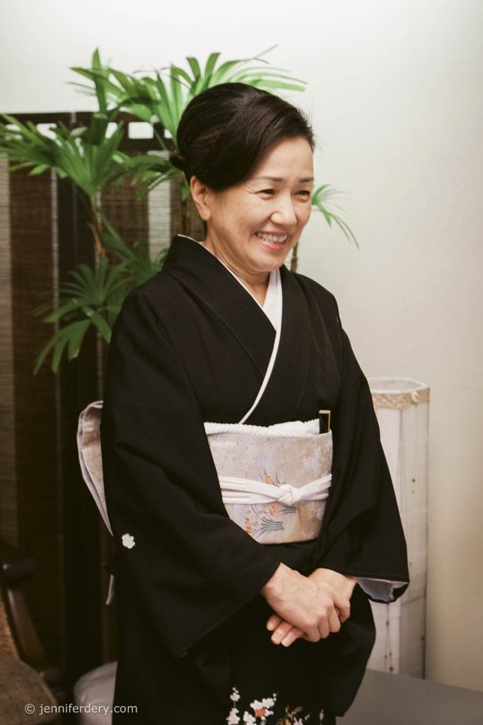 A woman wearing a traditional black kimono with a patterned obi smiles gently, standing indoors near a green potted plant and a wooden screen.