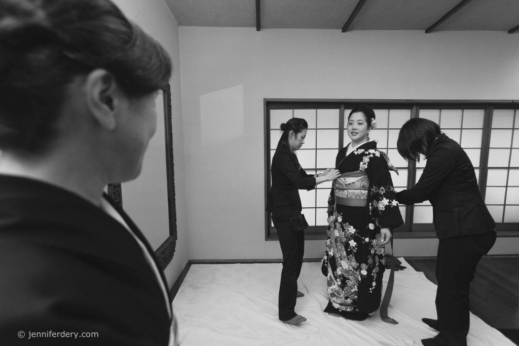 A woman in a black kimono stands as two attendants help adjust her outfit, while another woman looks on, smiling, in a traditional Japanese-style room with sliding shoji doors.