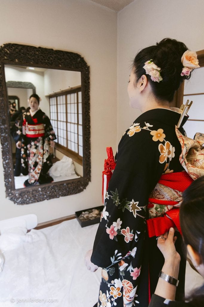 A woman wearing a black floral kimono and an ornate red obi stands in front of a mirror, while another person adjusts her obi. Her reflection shows her holding a decorative handbag and her hair adorned with flowers.