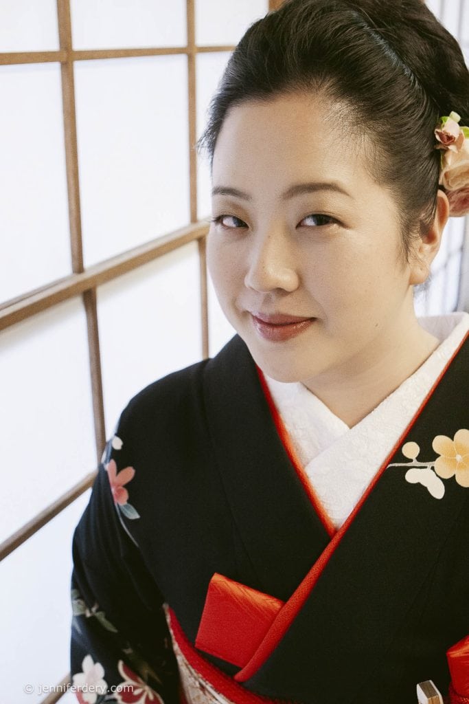A woman wearing a black floral kimono with red accents stands in front of a shoji screen, smiling gently. Her hair is neatly styled with a flower accessory.