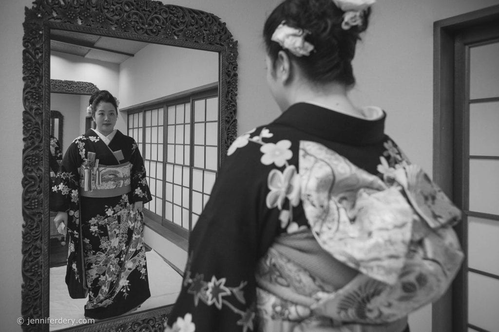 A woman wearing a traditional kimono stands in front of an ornate mirror, looking at her reflection in a room with shoji doors. The photo is in black and white.