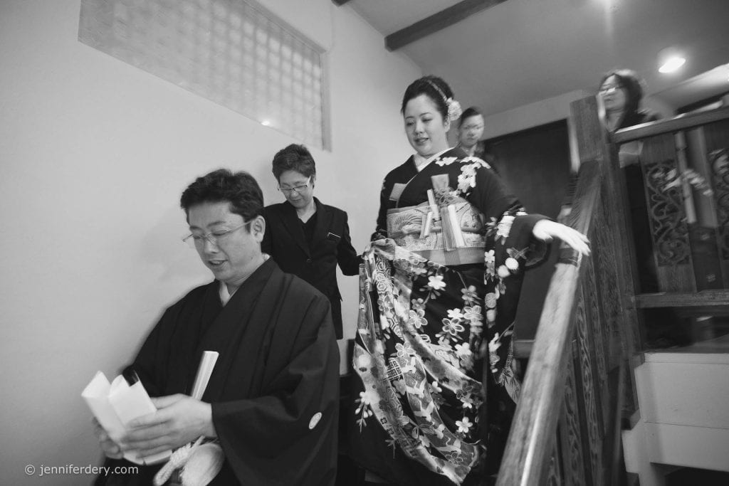 A group of people in traditional Japanese attire descend a wooden staircase, with a woman in a floral kimono at the center. The image is in black and white and captures a formal, possibly ceremonial moment.