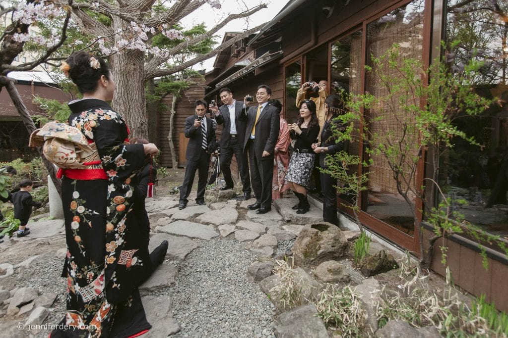 A woman in a black floral kimono faces a group of people dressed formally, posing for a photo outside a traditional wooden building surrounded by trees and plants.