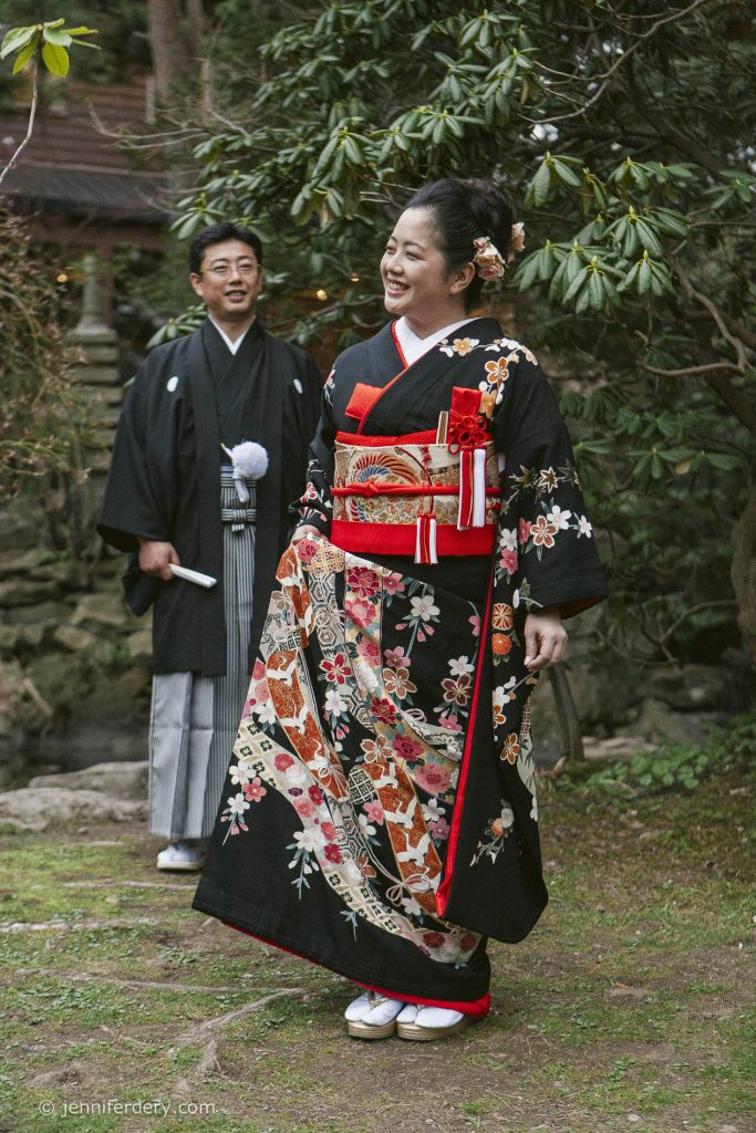 A woman in a black floral kimono and red obi smiles as she walks outdoors, while a man in a traditional black kimono stands behind her in a garden setting.