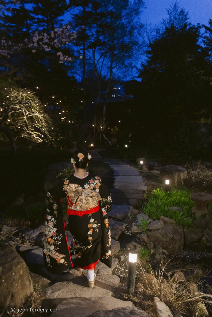 A person wearing a traditional black floral kimono walks along a lantern-lit stone path in a serene Japanese garden at dusk, surrounded by greenery and blooming trees.