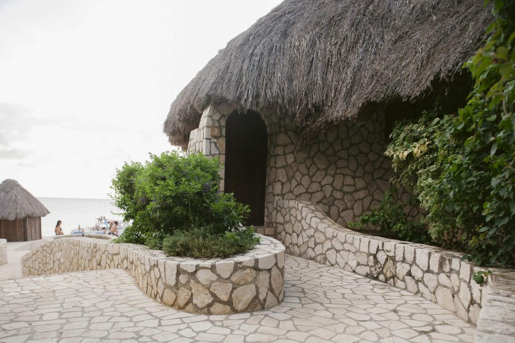Rockhouse Hotel, Negril, Jamaica: A stone building with a thatched roof sits near a beach, surrounded by greenery. There are curved stone paths and walls, and the ocean is visible in the background with people relaxing on the sand.
