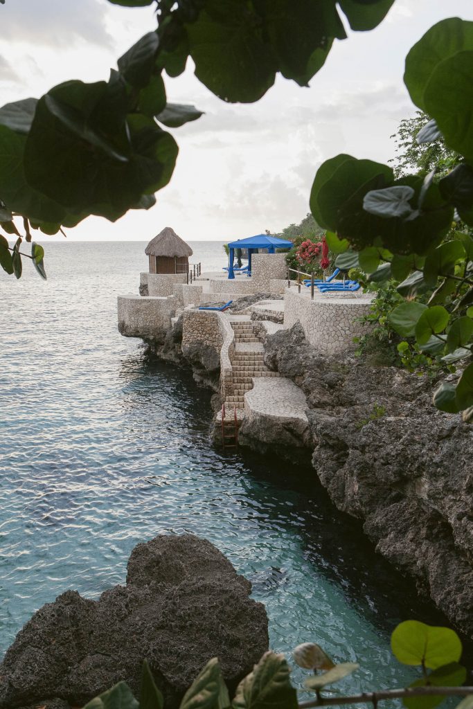 Rockhouse Hotel, Negril, Jamaica: A stone pathway and steps lead down to clear turquoise water from a rocky coastline, framed by leafy branches. Blue lounge chairs and a small thatched-roof hut sit atop the cliffs, overlooking the ocean.