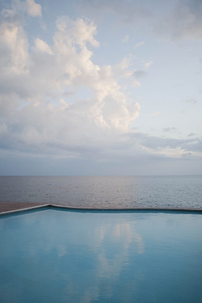 Rockhouse Hotel, Negril, Jamaica: A tranquil swimming pool overlooks a calm ocean under a cloudy sky, with soft sunlight reflecting on the water's surface.