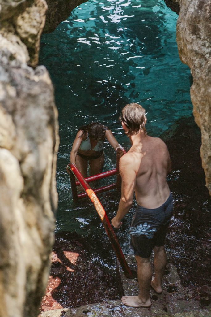A woman in a bikini climbs a red ladder from clear blue water surrounded by rocky cliffs, while a man in swim trunks stands at the top, watching her.