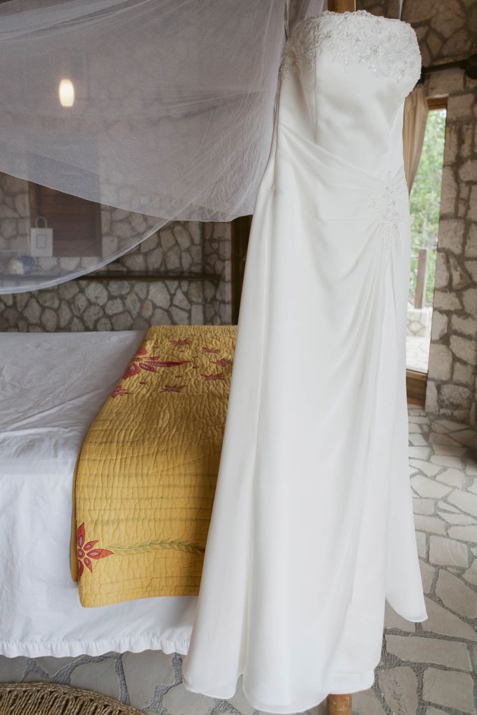 A white strapless wedding dress hangs on a bedpost in a rustic room with stone walls, a bed with a yellow quilt, and a mosquito net.