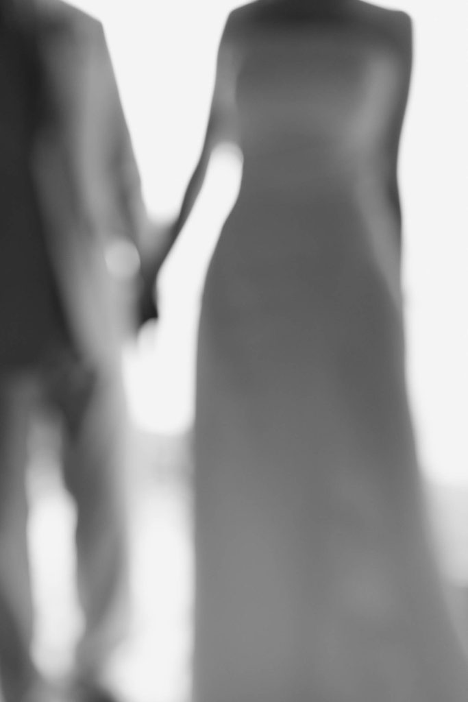 A blurred black-and-white image of two people holding hands, with one wearing a dress and the other in a suit, suggesting a formal or romantic occasion.