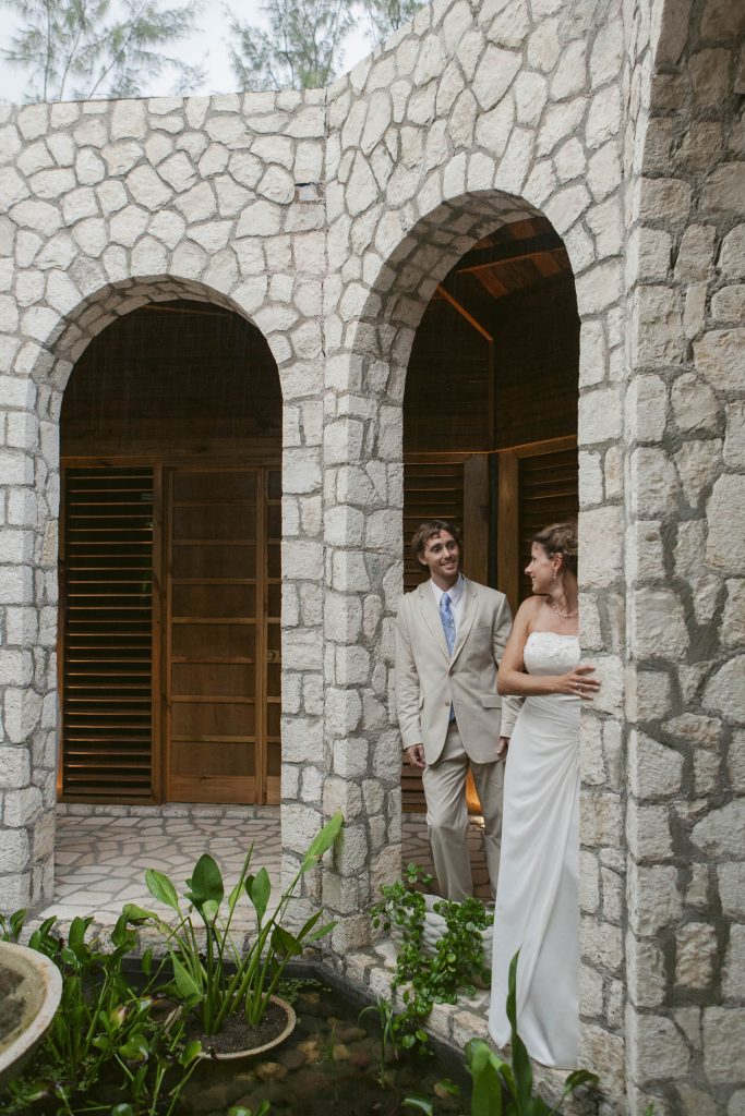 Rockhouse Hotel, Negril, Jamaica: A bride in a white dress stands by a stone archway, smiling at a groom in a light suit who approaches her. They are in a courtyard with greenery and a small pond, surrounded by stone walls and wooden doors.
