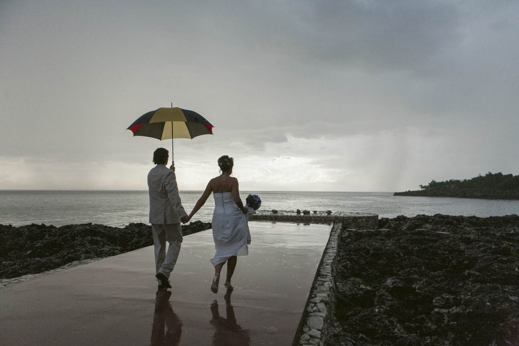 Rockhouse Hotel, Negril, Jamaica: A couple in formal attire walks hand in hand on a rocky path by the sea, sharing a red and yellow umbrella on a cloudy day. The woman holds a bouquet and the sky appears overcast with light in the distance.