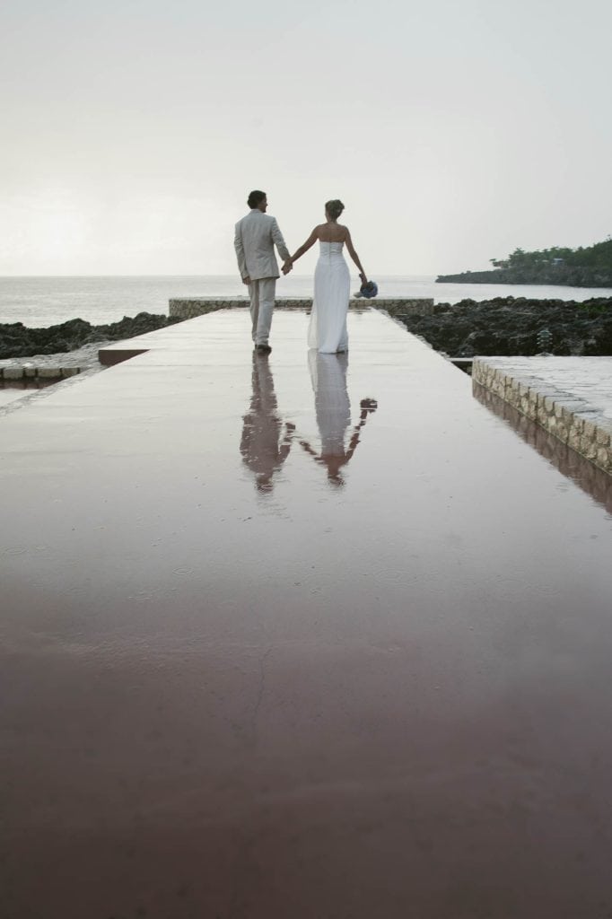 Rockhouse Hotel, Negril, Jamaica: A couple in wedding attire walks hand in hand on a wet pathway by the sea, their reflections visible on the shiny surface, with rocky shores and calm water in the background.