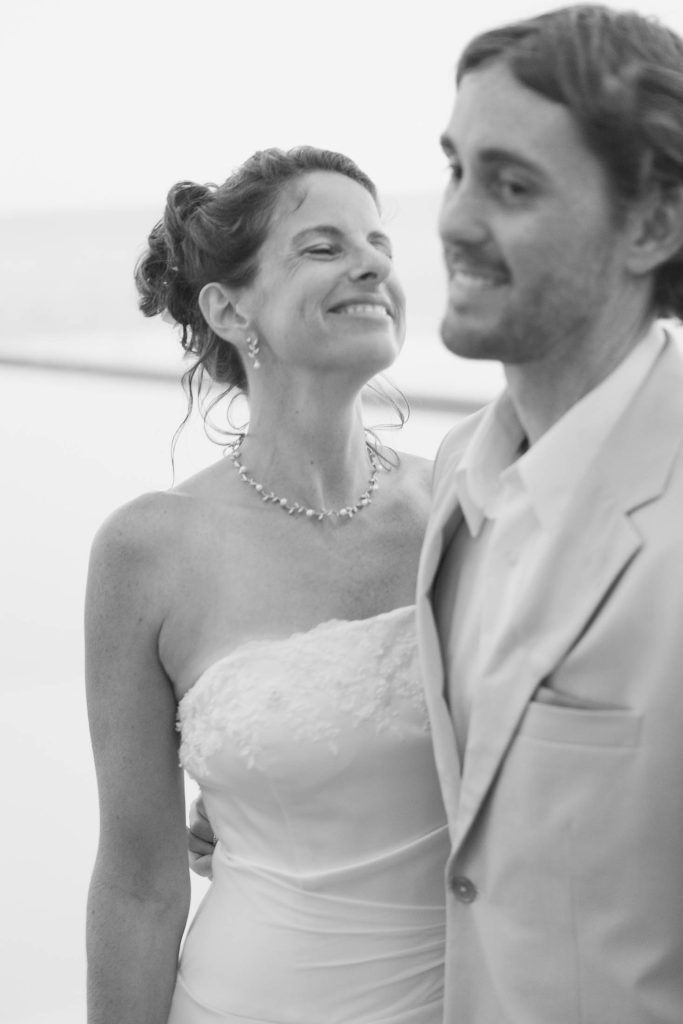 A bride in a strapless wedding dress smiles joyfully, standing next to a groom in a light-colored suit. The photo is black and white, with a soft, romantic feel.
