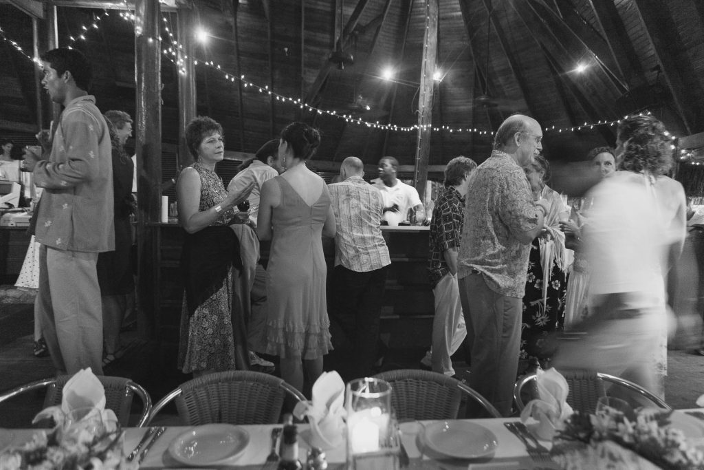 A group of people in semi-formal attire socialize around a bar inside a wooden structure decorated with string lights; a long table with set places is in the foreground. The scene is in black and white.