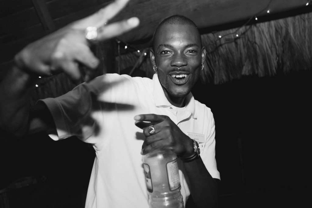 A man smiling and posing with a peace sign while holding a bottle. He wears a light-colored polo shirt and rings, and appears to be enjoying himself at a nighttime event with string lights in the background.
