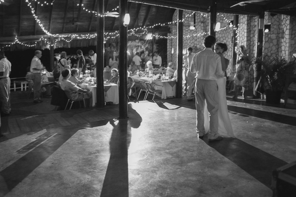A black-and-white photo of a wedding reception shows a couple dancing in the foreground, while guests sit at decorated tables and string lights hang from the ceiling, creating a warm and festive atmosphere.