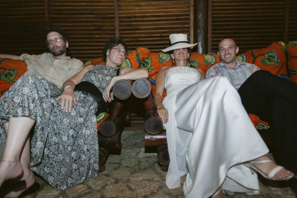 Four adults sit on a wooden couch with colorful patterned cushions. Two women in dresses and two men, one wearing a hat, relax together, all appearing comfortable and at ease in a warmly lit room with wood-paneled walls.