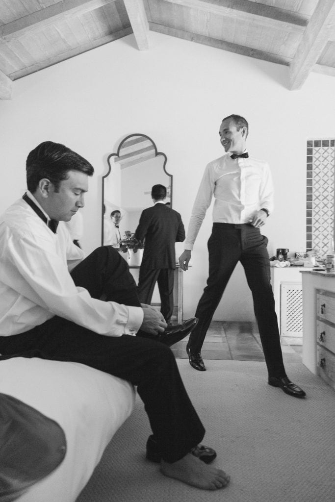 Two men in formal attire get ready in a bright room; one sits on a bed putting on his shoes, while the other stands smiling. A mirror reflects a third man in a suit in the background.