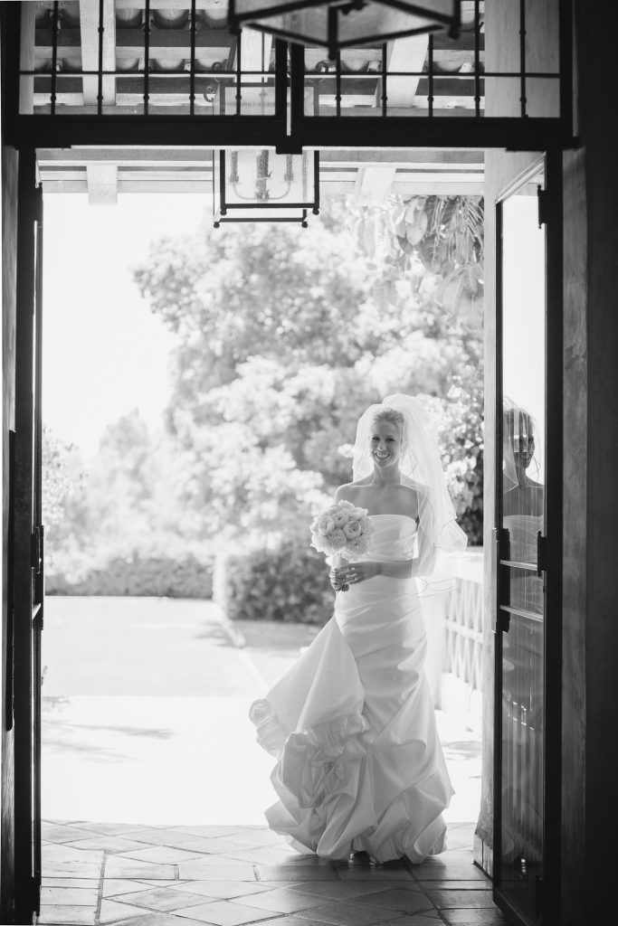 A bride in a strapless wedding gown and veil stands in a doorway, holding a bouquet of flowers, smiling and looking forward. Sunlight shines from outside, creating a bright, airy atmosphere.