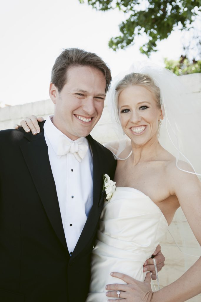 A smiling bride in a strapless white wedding dress and veil stands beside a smiling groom in a black tuxedo and white bow tie, posing outdoors in bright sunlight.