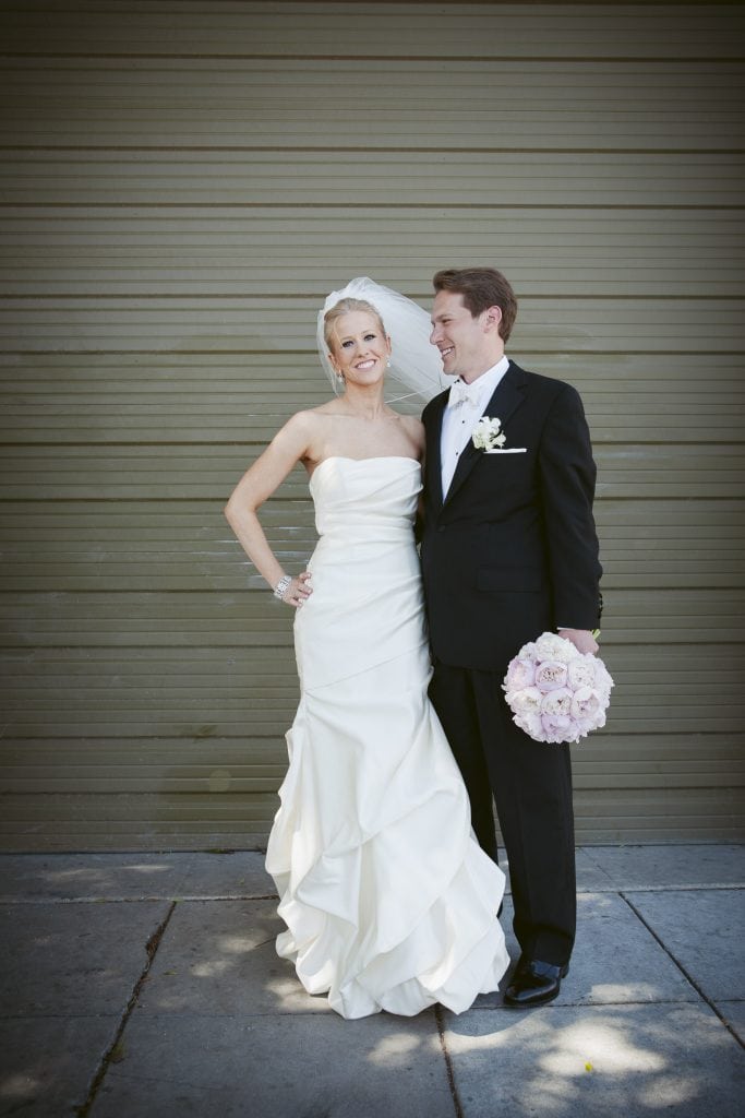 A bride in a strapless white gown and veil stands next to a groom in a black tuxedo holding a bouquet of pale pink flowers. They smile at each other in front of a green garage door.