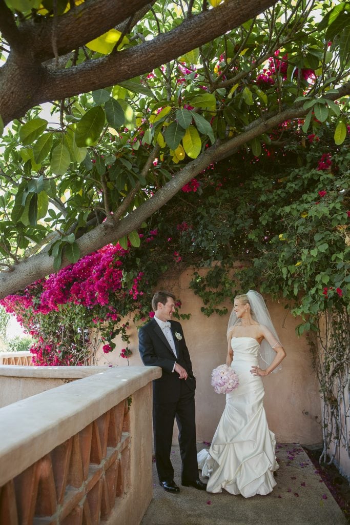 A bride in a white gown and veil holding a bouquet of pink flowers stands beside a groom in a black tuxedo under lush greenery and pink bougainvillea on a sunlit terrace.