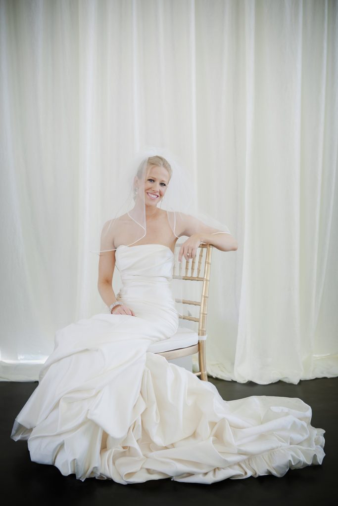 A bride in a strapless white wedding gown and veil sits sideways on a gold chair, smiling, with a soft white curtain backdrop behind her.