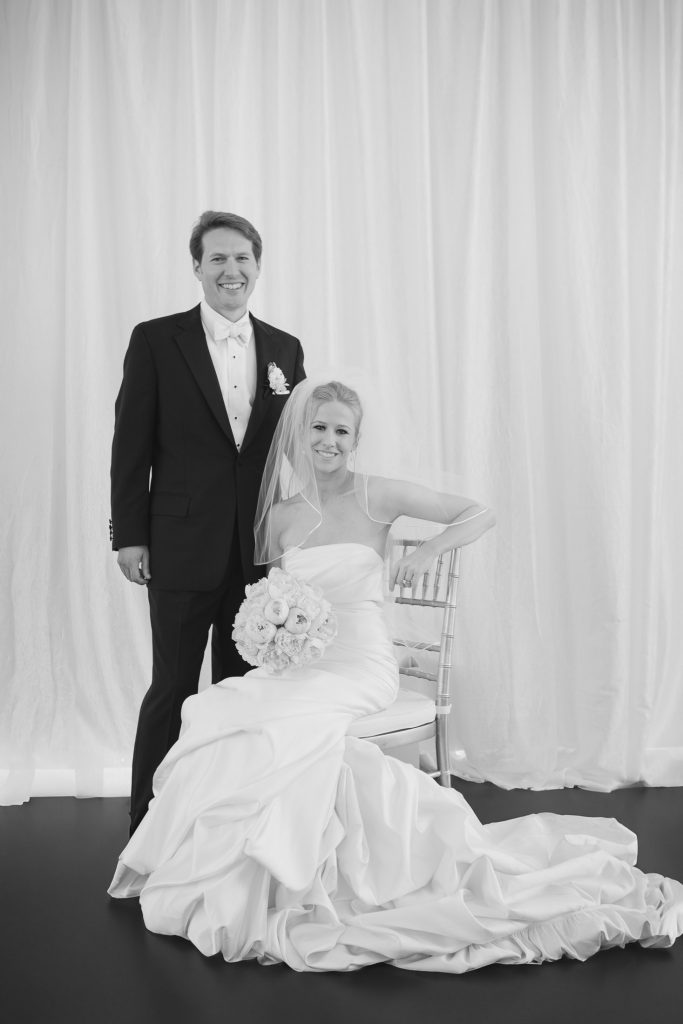 A smiling bride in a strapless gown and veil sits holding a bouquet of roses, while a groom in a black tuxedo stands beside her; both pose against a light, draped background.