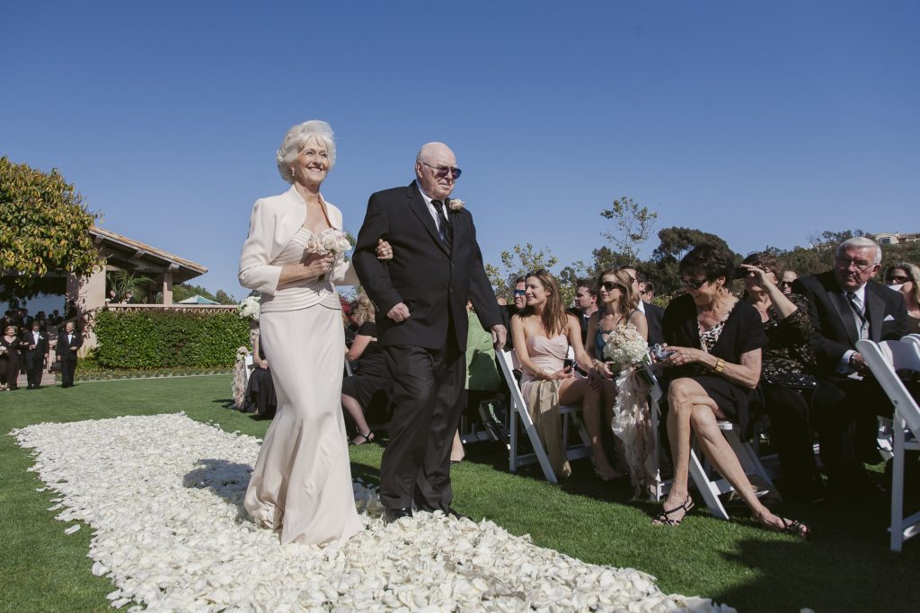 An elderly woman in a light suit and an elderly man in a black suit walk arm in arm down an outdoor aisle covered in white flower petals, surrounded by seated guests at a sunny wedding ceremony.