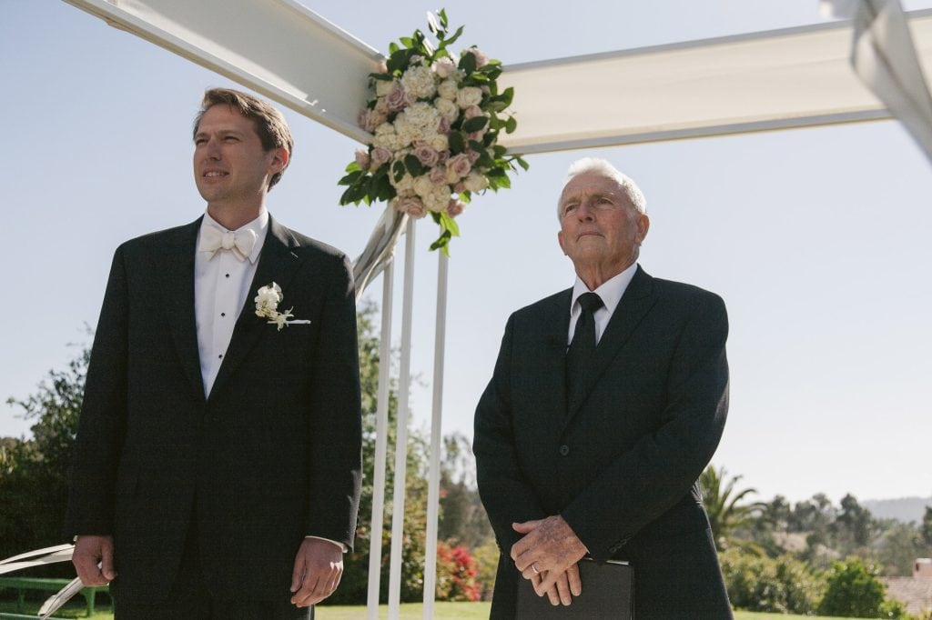 A groom and an older man in black suits stand under a floral wedding arch outdoors, looking forward. The sky is clear and sunny, with greenery and flowers visible in the background.