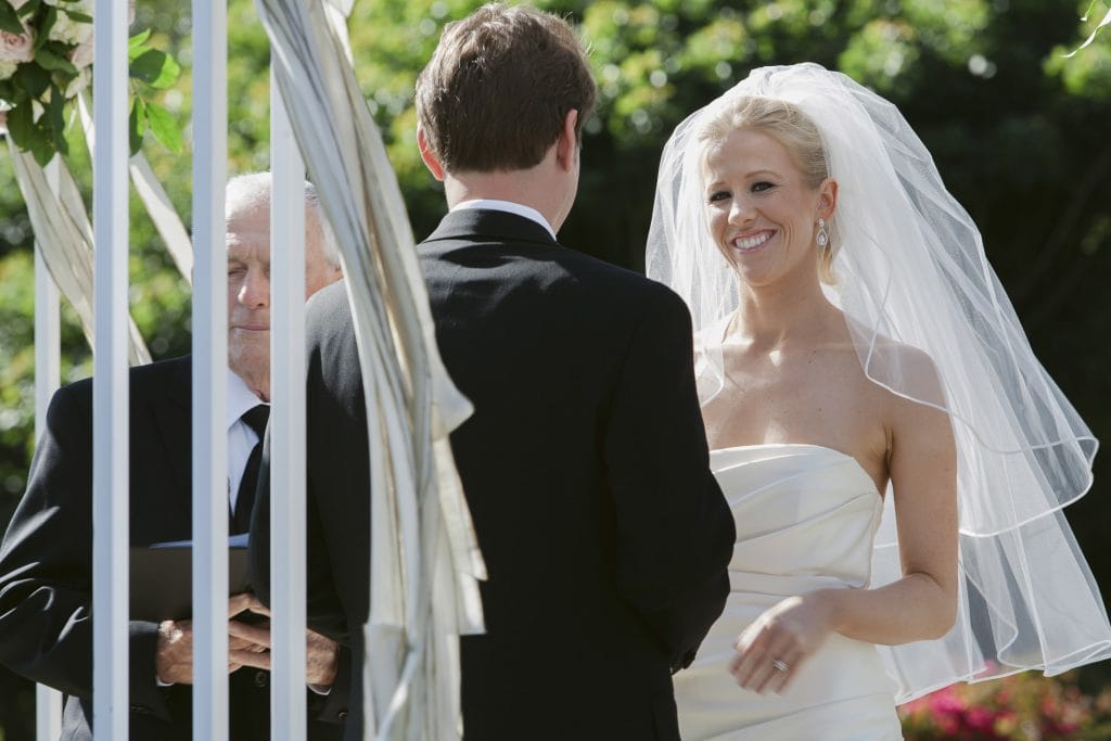 A bride in a white dress and veil smiles at the groom during an outdoor wedding ceremony, while an officiant stands beside them holding a book. Green foliage is visible in the background.