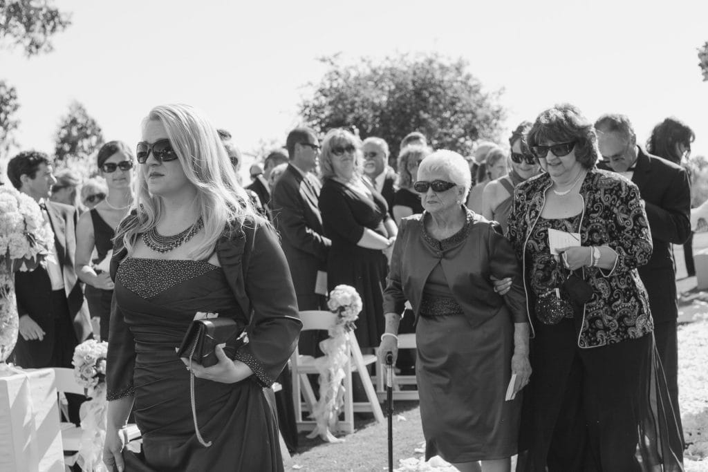 A group of formally dressed people, mostly women in dark dresses and sunglasses, walk together outdoors at what appears to be a wedding or formal event. Some people are seated in the background on benches.