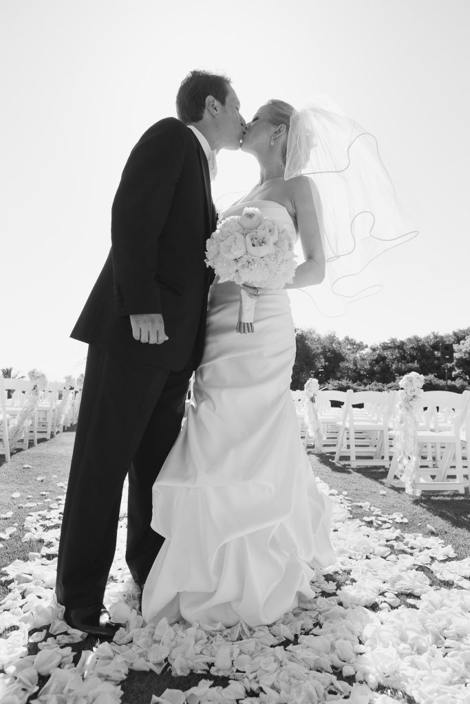 A bride and groom kiss outdoors, standing on a path covered with flower petals. The bride holds a bouquet and wears a long white dress and veil; the groom is in a dark suit. Rows of white chairs line the aisle behind them.