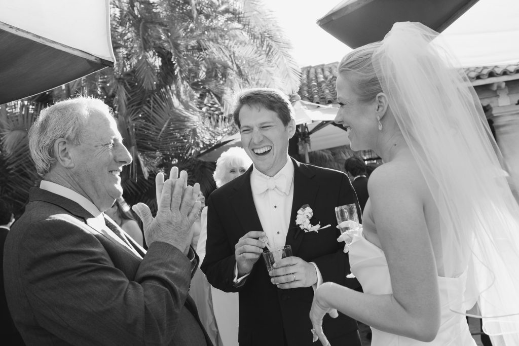 A bride and groom in wedding attire laugh joyfully with an older man at an outdoor reception, surrounded by palm trees and guests. The bride holds a champagne flute and wears a veil.