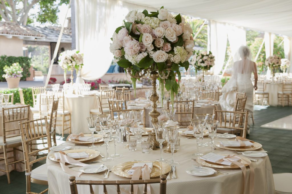 Elegant wedding reception table set with gold chairs, white linens, gold-rimmed plates, glassware, and a large centerpiece of white and pale pink flowers; a blurred bride stands in the background.