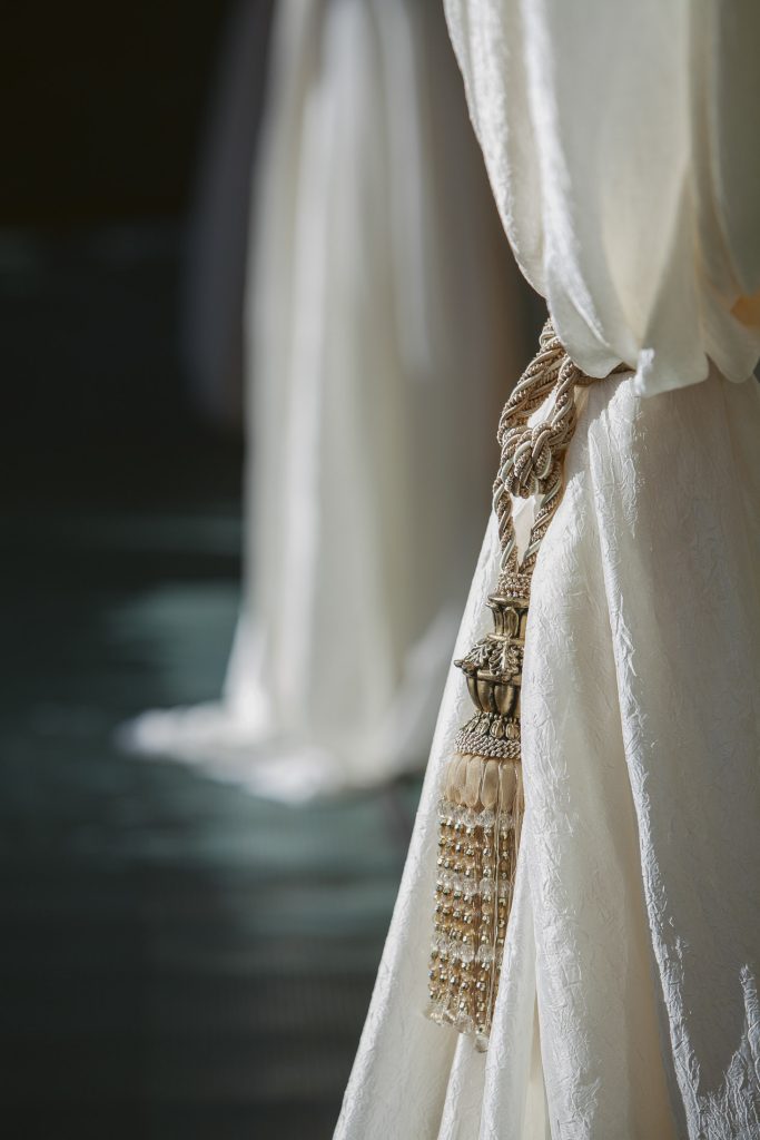 Close-up of an elegant cream-colored curtain tied back with an ornate braided rope and tassel, with soft sunlight highlighting its textured fabric; another curtain is blurred in the background.