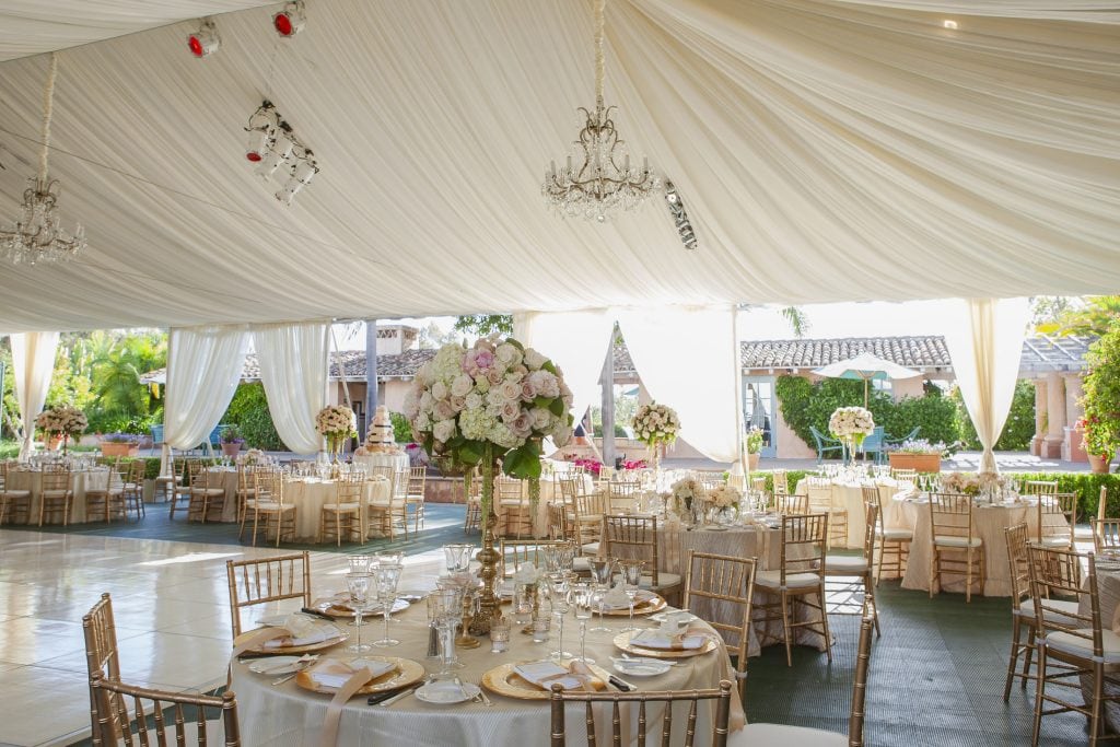 Elegant wedding reception setup under a white draped tent with chandeliers, round tables with gold chairs, floral centerpieces, and glassware, surrounded by lush greenery and sunlight streaming in.