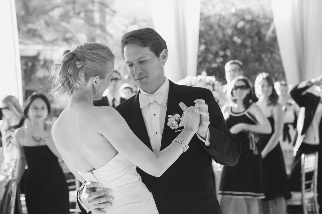 A bride and groom share their first dance at a wedding reception, surrounded by elegantly dressed guests watching and smiling. The scene is captured in black and white.