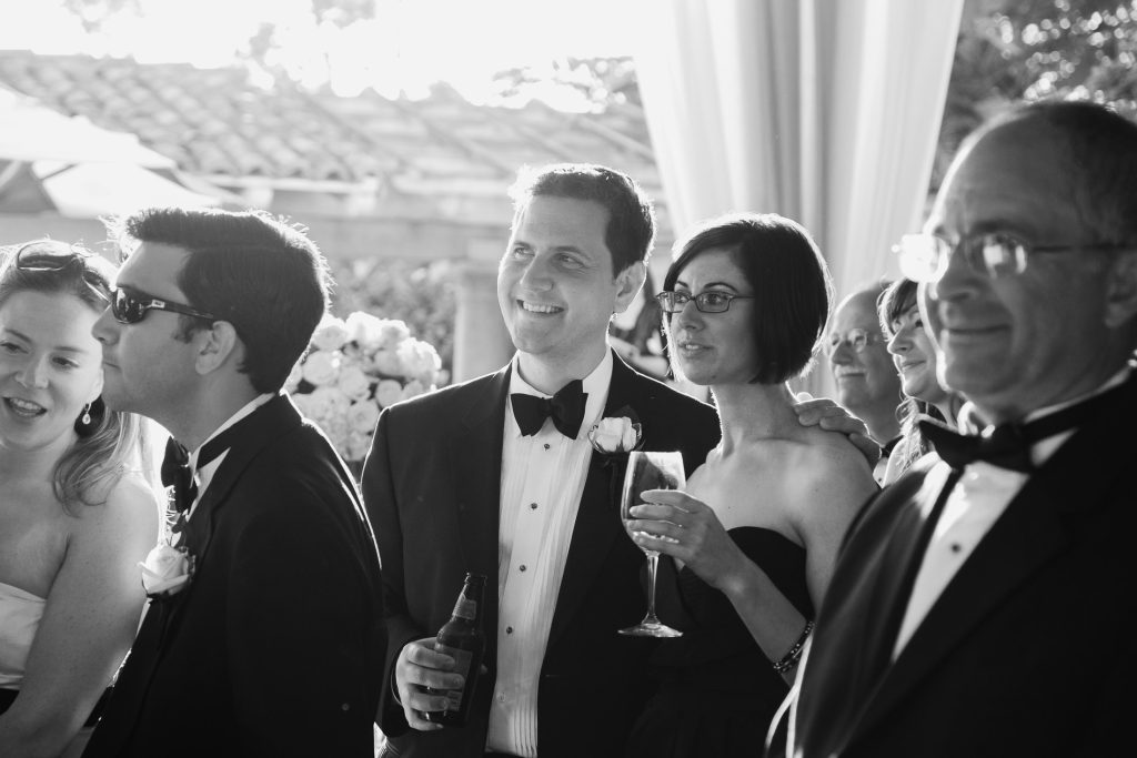 A group of people dressed formally at a wedding or formal event, smiling and standing together. One man holds a drink, and a woman beside him holds a champagne glass. The setting appears festive and celebratory.