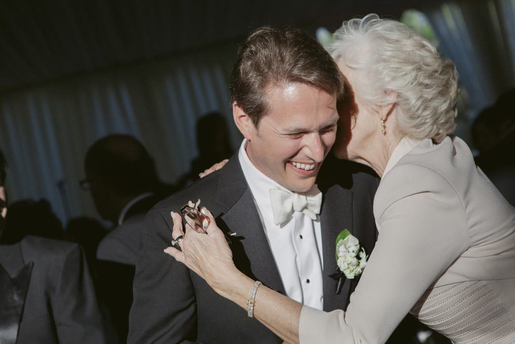 A woman with white hair hugs and whispers to a smiling man in a tuxedo at what appears to be a formal event or wedding reception.