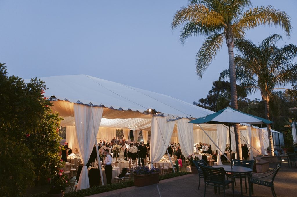 An outdoor wedding reception under a large white tent with draped fabric, elegant lighting, and guests dining at round tables. Palm trees and patio furniture surrounded the festive evening scene.
