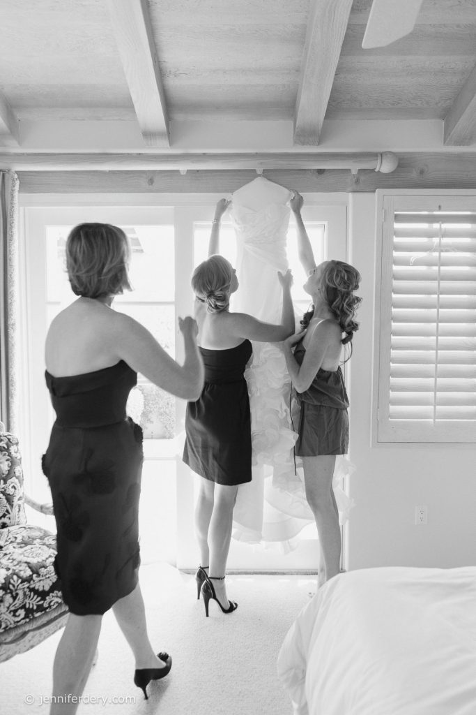 Three women in a bright room help prepare a wedding dress, with one holding it up by the shoulders. Sunlight streams through large windows and shutters, creating a cheerful, airy atmosphere.